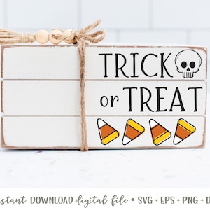 Puede incluir: Un letrero de madera blanco con el texto "Trick or Treat" en letras negras. Un icono de calavera está al lado de la palabra "Trick". Cinco iconos de maíz dulce están debajo del texto.