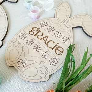 Op de afbeelding: Houten paasei-knutselwerk met konijnenoren, klaar om te worden beschilderd. Het ei heeft een golvend lijnenontwerp en de naam "Gracie" is aan de voorkant gegraveerd.