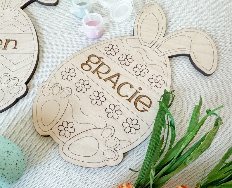Op de afbeelding: Houten paasei-knutselwerk met konijnenoren, klaar om te worden beschilderd. Het ei heeft een golvend lijnenontwerp en de naam "Gracie" is aan de voorkant gegraveerd.