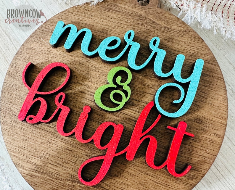 May include: A wooden round sign with the words "merry & bright" in red, blue, and green paint.