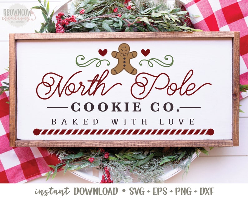 May include: A white sign with a brown wooden frame, featuring a gingerbread man cookie, red hearts, and the text "North Pole - COOKIE CO. - BAKED WITH LOVE".