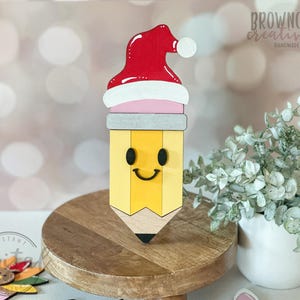 May include: A wooden pencil-shaped decoration with a Santa hat. The pencil is yellow with a smiling face and a black tip. The Santa hat is red with a white trim and a white pom-pom. The decoration is on a wooden stand.