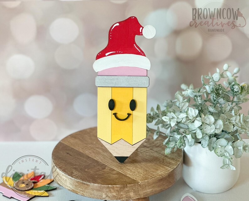 May include: A wooden pencil-shaped decoration with a Santa hat. The pencil is yellow with a smiling face and a black tip. The Santa hat is red with a white trim and a white pom-pom. The decoration is on a wooden stand.