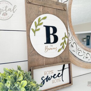 May include: A wooden sign with a white circle in the center. The circle has a black letter "B" and the text "The Brownings" in gold script. The sign also has a smaller sign below it that says "Home Sweet Home" in black script.