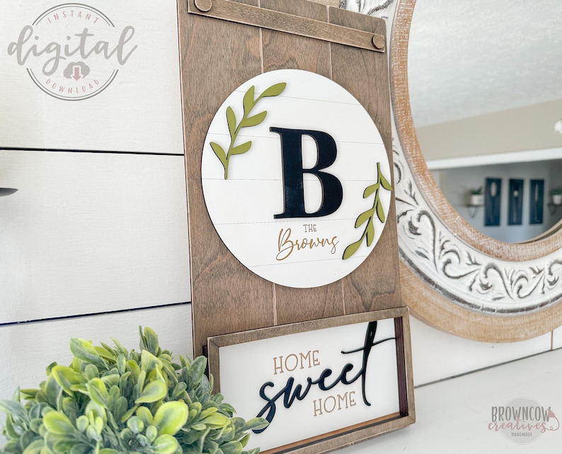 May include: A wooden sign with a white circle in the center. The circle has a black letter "B" and the text "The Brownings" in gold script. The sign also has a smaller sign below it that says "Home Sweet Home" in black script.