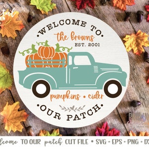 Pode incluir: Um sinal redondo branco com o texto "Welcome to the Browns Est. 2001 pumpkins. cider •Our Patch." Um caminhão azul-verde com uma caçamba cheia de abóboras laranja está no centro do sinal.