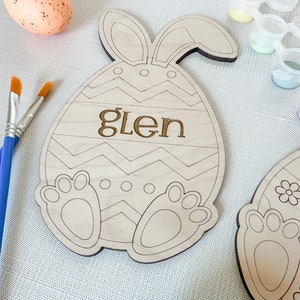 Op de afbeelding: Een houten paasei-vormig knutselwerk met konijnenoren, een zigzagpatroon en de naam "Glen" erop. Het knutselwerk is klaar om te worden geschilderd met de meegeleverde verf.