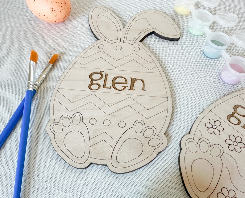 Op de afbeelding: Een houten paasei-vormig knutselwerk met konijnenoren, een zigzagpatroon en de naam "Glen" erop. Het knutselwerk is klaar om te worden geschilderd met de meegeleverde verf.