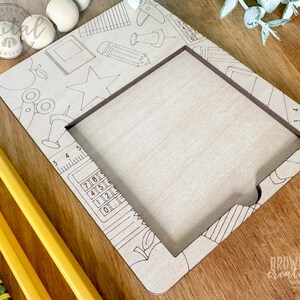 May include: A wooden frame with a cutout for a photo or artwork. The frame is decorated with a school-themed design, including a ruler, a calculator, a pencil, a star, and a scissors. The frame is unfinished and ready to be painted or stained.