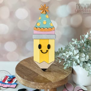 May include: A wooden pencil-shaped decoration with a smiling face and a party hat. The pencil is yellow with a black tip, and the hat is teal with yellow polka dots. The decoration is on a wooden stand. The text "Browncow Creatives Handmade" is visible.