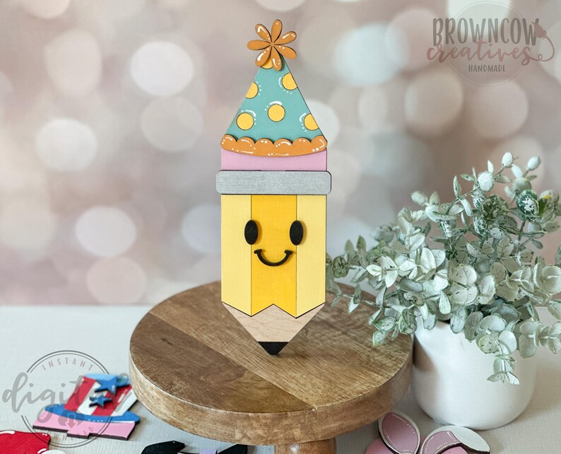 May include: A wooden pencil-shaped decoration with a smiling face and a party hat. The pencil is yellow with a black tip, and the hat is teal with yellow polka dots. The decoration is on a wooden stand. The text "Browncow Creatives Handmade" is visible.