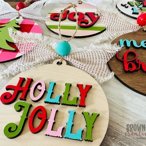 May include: A set of five wooden Christmas ornaments with various holiday greetings. The ornaments are painted in red, green, pink, blue, and white with black lettering. The ornaments are decorated with burlap ribbon and a small wooden bead.