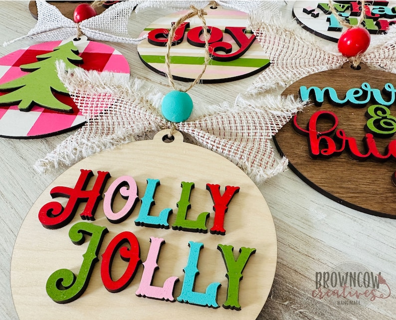 May include: A set of five wooden Christmas ornaments with various holiday greetings. The ornaments are painted in red, green, pink, blue, and white with black lettering. The ornaments are decorated with burlap ribbon and a small wooden bead.