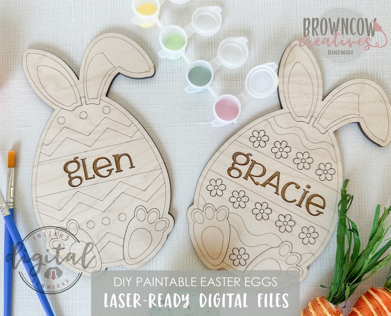 Op de afbeelding: Twee onafgewerkte houten paaseieren met konijnenoren. De eieren zijn versierd met verschillende patronen en hebben de namen "glen" en "gracie" gegraveerd. De eieren zijn klaar om te worden beschilderd.