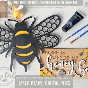 Puede incluir: Diseño de abeja de miel de madera cortada con láser con alas de panal y un cuerpo rayado amarillo y negro. La abeja es parte de un conjunto para un mini-apoyo intercambiable de madera. El letrero dice "Home is where my honey bee".