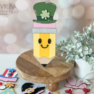 May include: A wooden pencil-shaped decoration with a smiling face, topped with a green leprechaun hat and shamrock. The pencil is yellow with a gray band and pink accent. The pencil is on a wooden stand with other holiday-themed decorations.