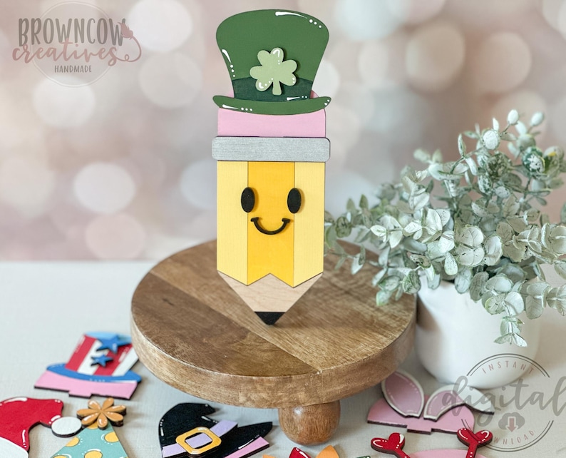May include: A wooden pencil-shaped decoration with a smiling face, topped with a green leprechaun hat and shamrock. The pencil is yellow with a gray band and pink accent. The pencil is on a wooden stand with other holiday-themed decorations.