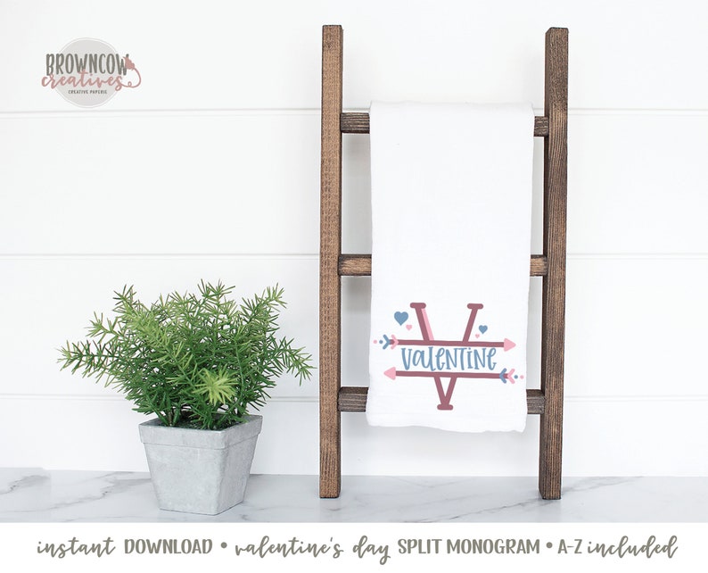 May include: White Valentine's Day split monogram towel with a brown wooden ladder. The towel features a large letter "V" with the word "VALENTINE" and arrow accents in pink and blue. A small potted plant is on the left.