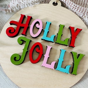 May include: A round wooden ornament with the words "HOLLY JOLLY" in colorful letters. The letters are cut out of wood and painted in red, green, pink, and blue.