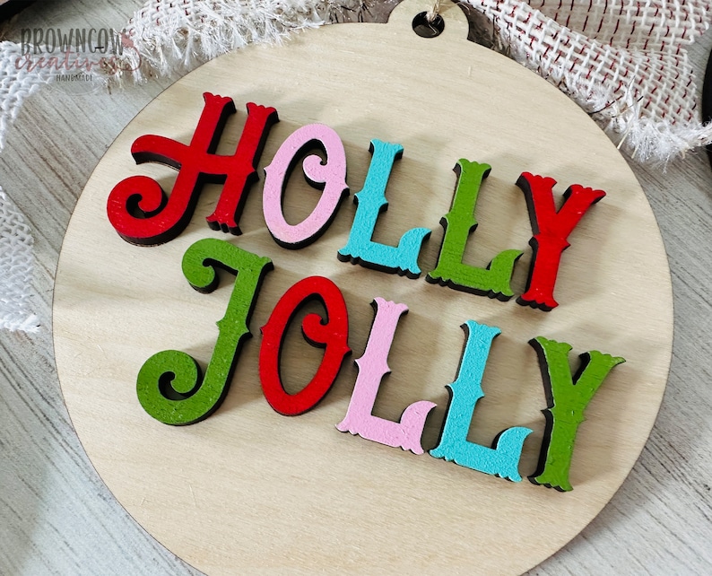 May include: A round wooden ornament with the words "HOLLY JOLLY" in colorful letters. The letters are cut out of wood and painted in red, green, pink, and blue.