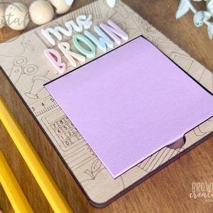 May include: A wooden teacher desk sign with the words "Mrs. Brown" in colorful letters. The sign has a built-in notepad and a ruler with a calculator design.