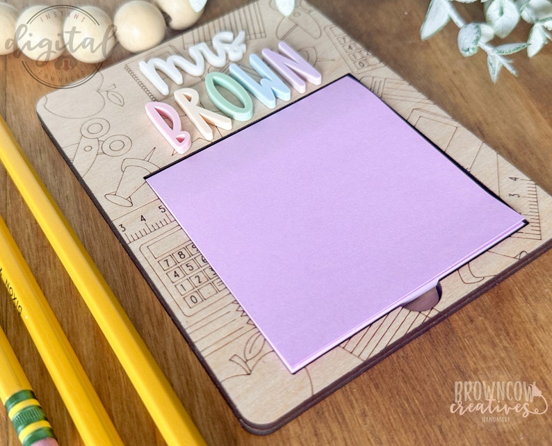 May include: A wooden teacher desk sign with the words "Mrs. Brown" in colorful letters. The sign has a built-in notepad and a ruler with a calculator design.