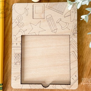May include: A wooden frame with a cutout for a photo or picture. The frame is decorated with a school-themed design, including a pencil, ruler, calculator, and apple. The text "BROWN COW CREATIVES" is visible in the bottom right corner.