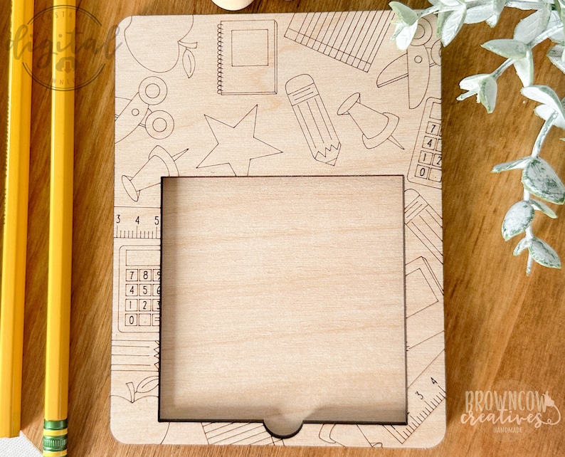 May include: A wooden frame with a cutout for a photo or picture. The frame is decorated with a school-themed design, including a pencil, ruler, calculator, and apple. The text "BROWN COW CREATIVES" is visible in the bottom right corner.