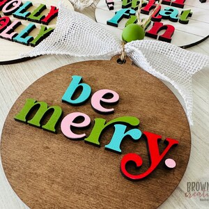 May include: A wooden round ornament with the words "be merry" in colorful letters. The letters are in shades of green, pink, blue, and red. The ornament is on a white wooden background.