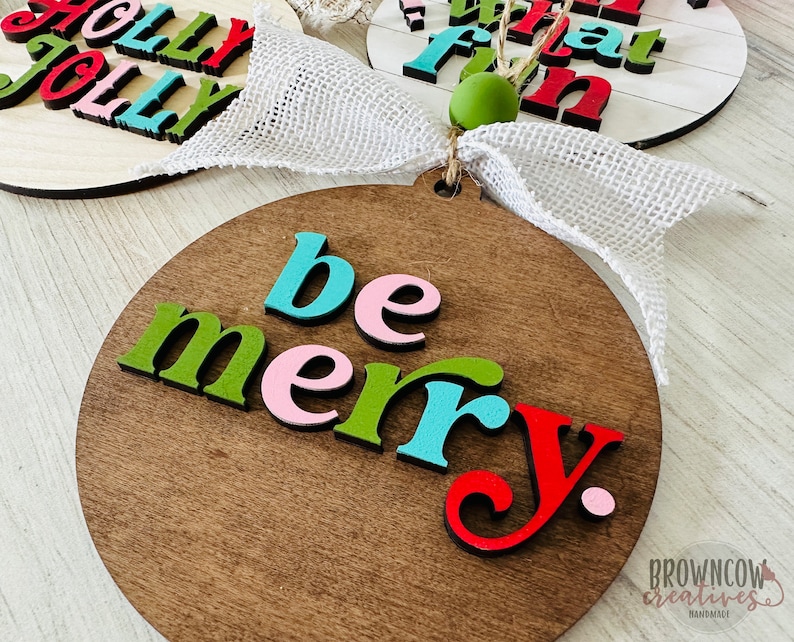 May include: A wooden round ornament with the words "be merry" in colorful letters. The letters are in shades of green, pink, blue, and red. The ornament is on a white wooden background.
