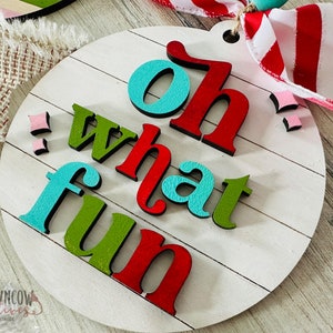 May include: A round wooden sign with the words "Oh what fun" in colorful letters. The letters are raised and have a 3D effect. The sign is painted white with a wood grain texture.