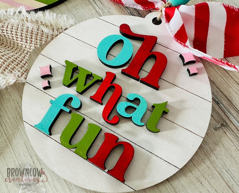 May include: A round wooden sign with the words "Oh what fun" in colorful letters. The letters are raised and have a 3D effect. The sign is painted white with a wood grain texture.