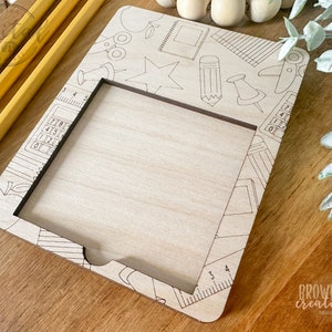May include: A wooden frame with a cut-out rectangle in the center. The frame is decorated with a school-themed design, including a pencil, ruler, calculator, and apple. The frame is perfect for displaying a photo or artwork.