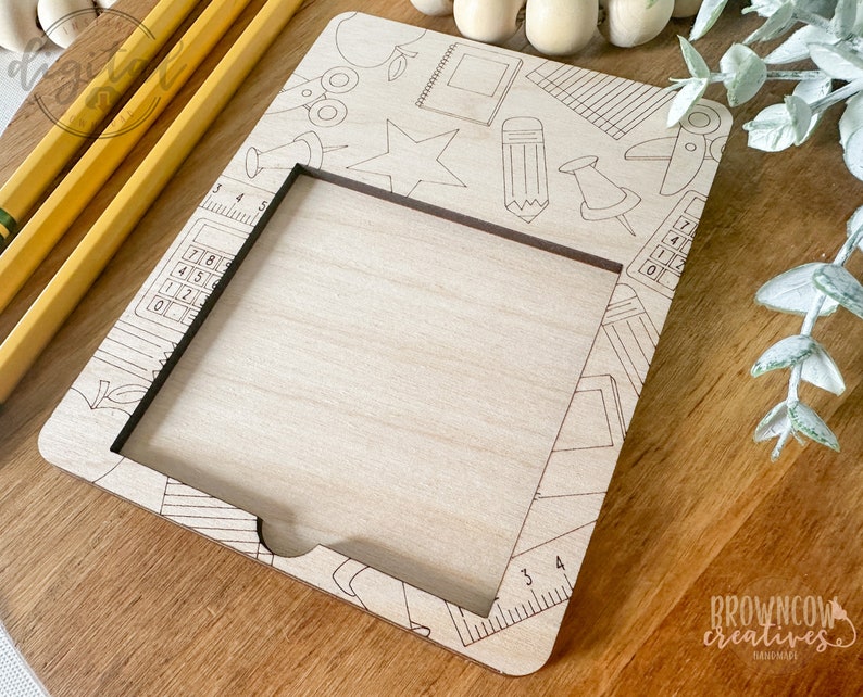 May include: A wooden frame with a cut-out rectangle in the center. The frame is decorated with a school-themed design, including a pencil, ruler, calculator, and apple. The frame is perfect for displaying a photo or artwork.