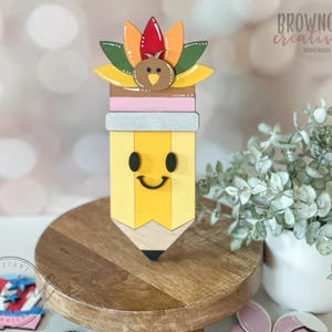 May include: A wooden pencil-shaped decoration with a smiling face, topped with a turkey and colorful feathers. The pencil is yellow with a wooden tip and a pink and gray band. The decoration is on a wooden stand.
