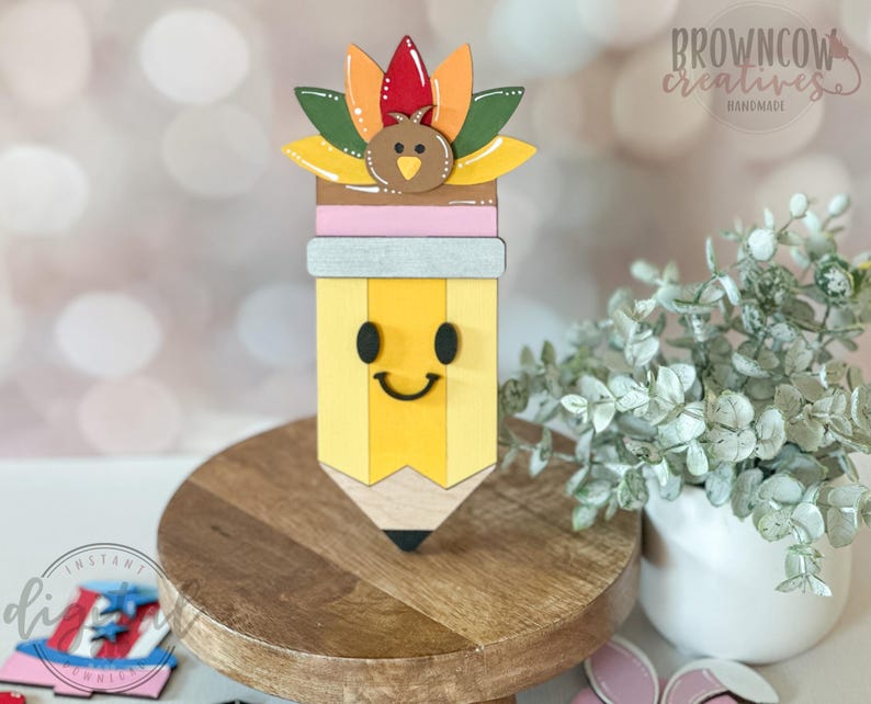 May include: A wooden pencil-shaped decoration with a smiling face, topped with a turkey and colorful feathers. The pencil is yellow with a wooden tip and a pink and gray band. The decoration is on a wooden stand.