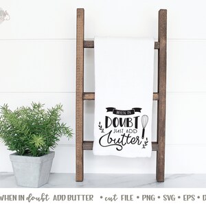 K&ouml;nnte beinhalten: Ein wei&szlig;es K&uuml;chentuch mit schwarzem Text, der "When in doubt, just add butter!" lautet, mit einer Grafik eines Schneebesens. Das Tuch h&auml;ngt an einer Holzleiter.