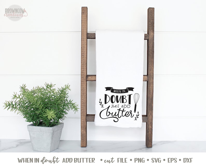 K&ouml;nnte beinhalten: Ein wei&szlig;es K&uuml;chentuch mit schwarzem Text, der "When in doubt, just add butter!" lautet, mit einer Grafik eines Schneebesens. Das Tuch h&auml;ngt an einer Holzleiter.