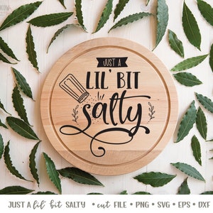 K&ouml;nnte beinhalten: Ein rundes, h&ouml;lzernes Schneidebrett mit dem Text "Just a Lil' Bit Salty" in schwarzer Schrift. Neben dem Text ist ein kleiner Salzstreuer abgebildet.