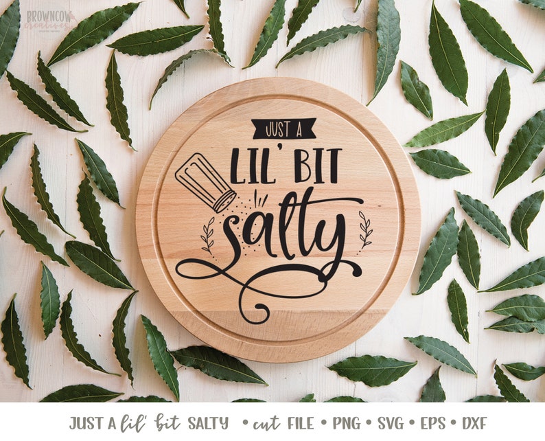 K&ouml;nnte beinhalten: Ein rundes, h&ouml;lzernes Schneidebrett mit dem Text "Just a Lil' Bit Salty" in schwarzer Schrift. Neben dem Text ist ein kleiner Salzstreuer abgebildet.