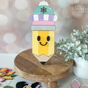 May include: A wooden pencil-shaped decoration with a smiling face and a winter hat. The pencil is yellow with a black tip and a gray eraser. The hat is light blue, purple, and white with a snowflake. The Browncow Creatives logo is visible.
