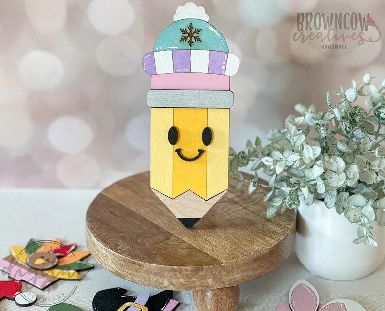May include: A wooden pencil-shaped decoration with a smiling face and a winter hat. The pencil is yellow with a black tip and a gray eraser. The hat is light blue, purple, and white with a snowflake. The Browncow Creatives logo is visible.