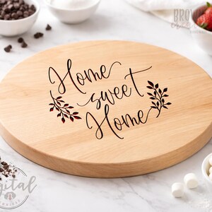 May include: A round wooden serving board with the phrase "Home sweet Home" in elegant script, framed by leaf accents. The board is light-colored and appears to be made of wood. The board is surrounded by bowls of ingredients, including chocolate chips, strawberries, and marshmallows.