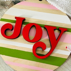 May include: A round wooden sign with a striped pattern in pink, white, beige, and green. The word "JOY" is written in red, stylized letters.