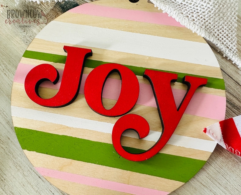 May include: A round wooden sign with a striped pattern in pink, white, beige, and green. The word "JOY" is written in red, stylized letters.