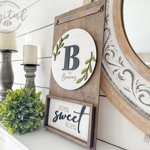 May include: A wooden sign with a white circle and the letter "B" in black with green leaves around it. The text "The Browns" is written in cursive below the letter. A smaller wooden sign below says "Home Sweet Home" in black cursive lettering.