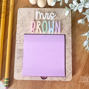 May include: A wooden note holder with a pink sticky note and the text "mrs BROWN" in colorful letters. The holder has a school-themed design with images of pencils, scissors, and a calculator.
