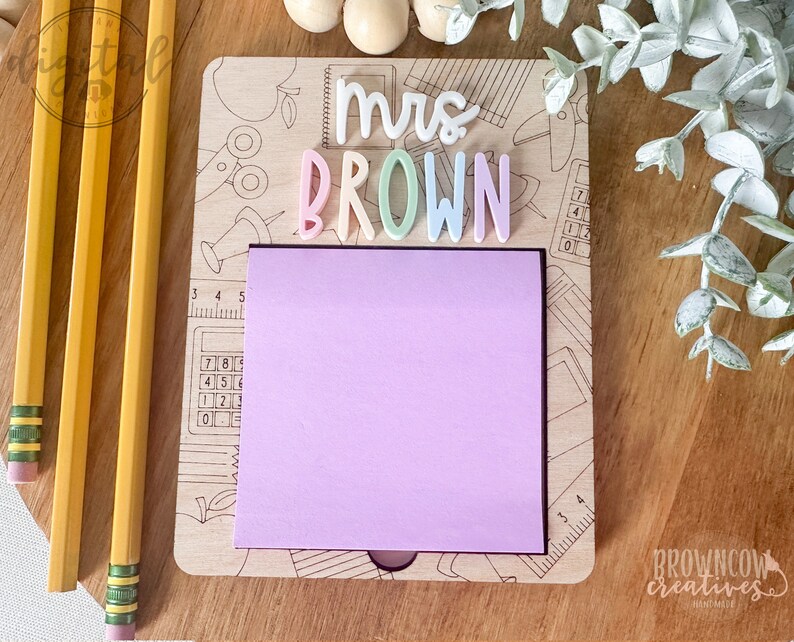 May include: A wooden note holder with a pink sticky note and the text "mrs BROWN" in colorful letters. The holder has a school-themed design with images of pencils, scissors, and a calculator.