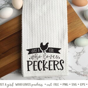 K&ouml;nnte beinhalten: Ein wei&szlig;es K&uuml;chentuch mit schwarzem Text, der "Just a girl who loves peckers" lautet. Eine schwarze Silhouette eines Huhns befindet sich &uuml;ber dem Text. Das Tuch liegt auf einer Holzoberfl&auml;che mit Eiern.