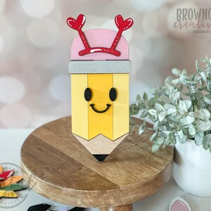 May include: A wooden pencil-shaped decoration with a smiling face, yellow body, and black tip. It has a pink top with red heart-shaped accents and the text "browncow creatives handmade". The pencil sits on a wooden stand.